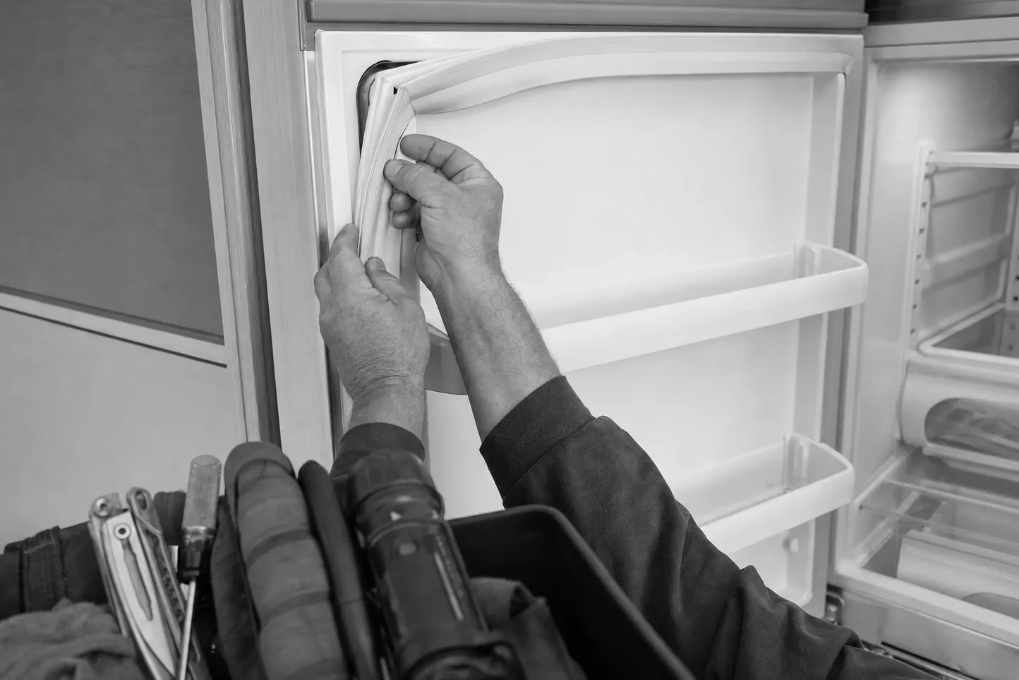 Black-and-white frame of two hands easing a door gasket out of its channel at the top corner of an open refrigerator door