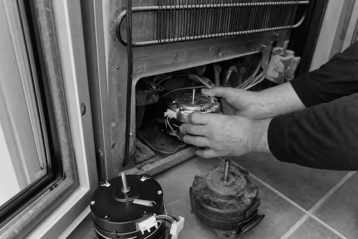 Black-and-white frame of hands lifting a fan motor into the lower bay of a cabinet, the corroded old motor standing on the tiles
